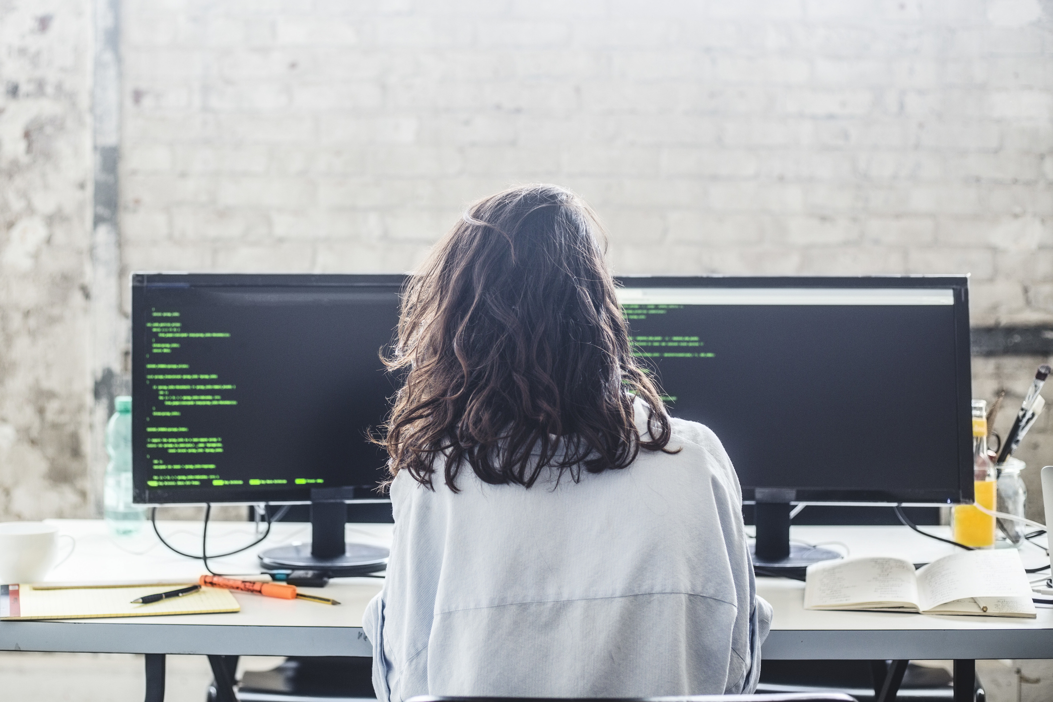 back of woman sitting at desk with two monitors