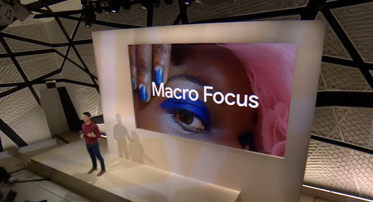 A speaker on the Google stage from Made by Google Event 2022, with the words Macro Focus on a screen behind them