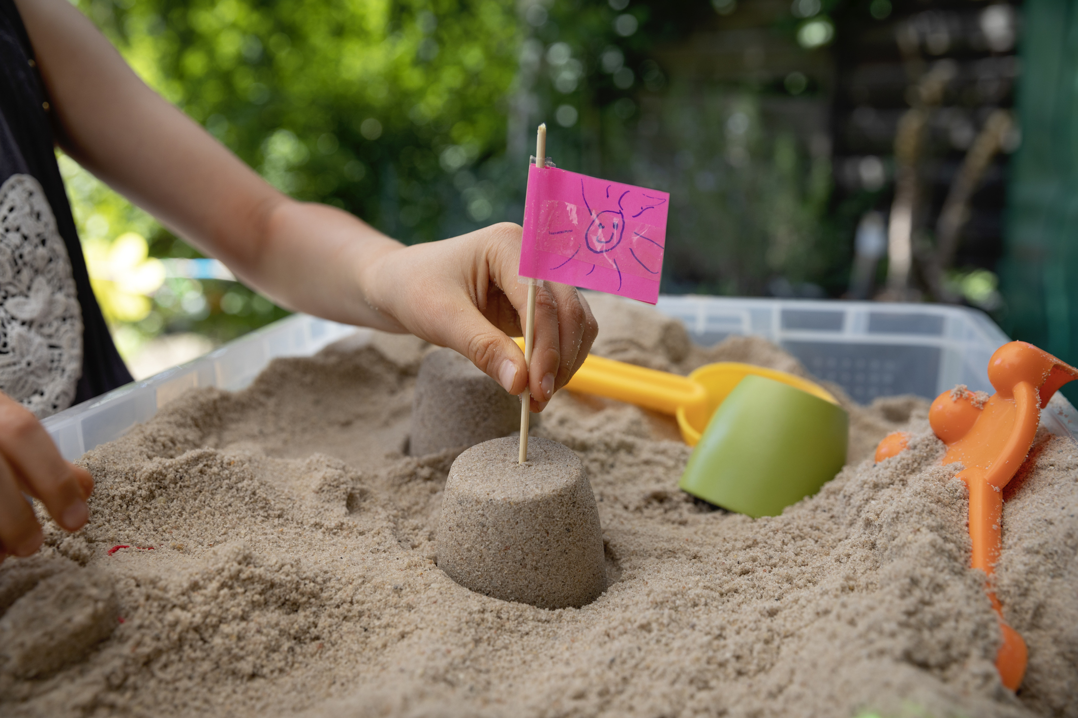 Child placing a homemade flag made from a pink sticky note with a drawing of a smiley face, into the top of a small sandcastle surrounded by sand inside a plastic sandbox.