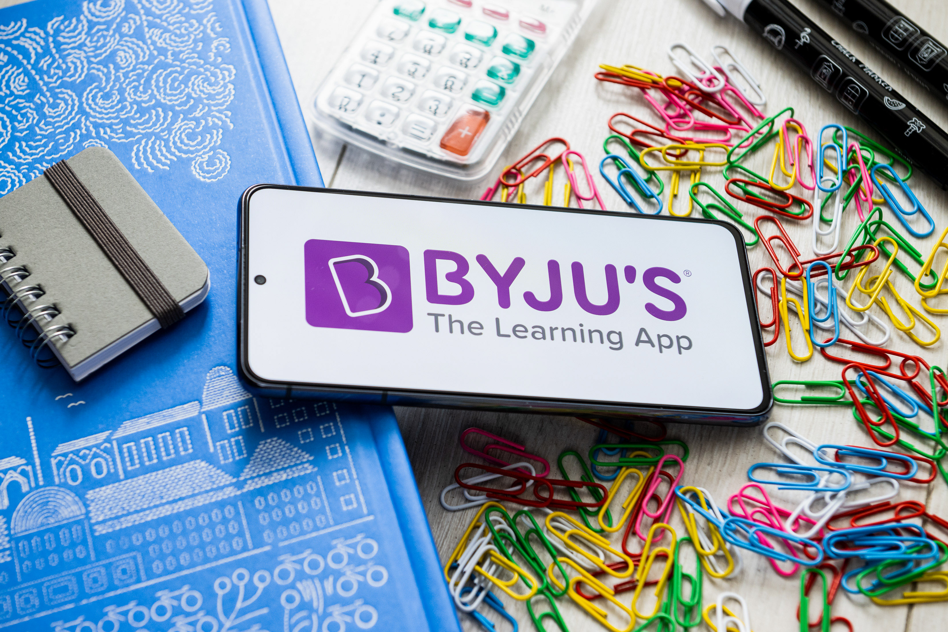 Byju's logo displayed on a smartphone laying on a table covered in school supplies
