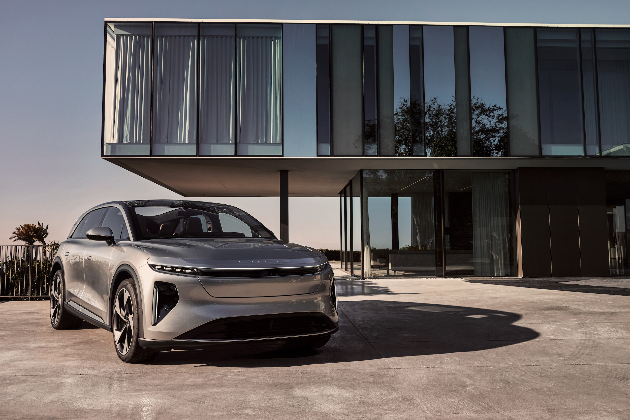 A promotional shot shows a silver Lucid Gravity SUV parked in front of a modern house with big glass windows.