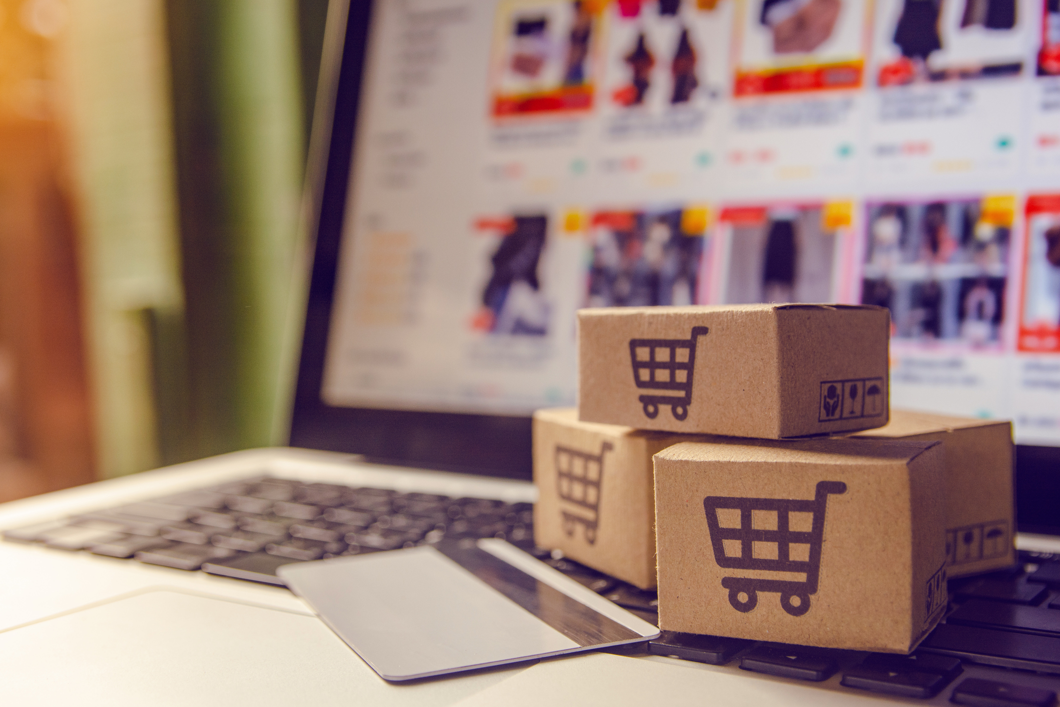 A stack of three cardboards boxes with the image of a shopping cart printed on them, resting on top of a laptop with the screen open to an e-commerce marketplace