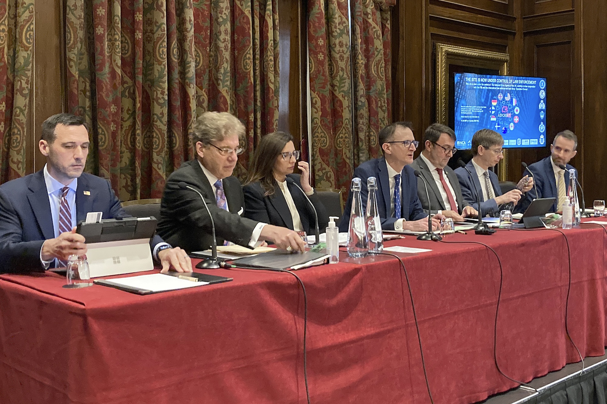 a photo of UK and US officials sitting at a red table discussing the LockBit takedown