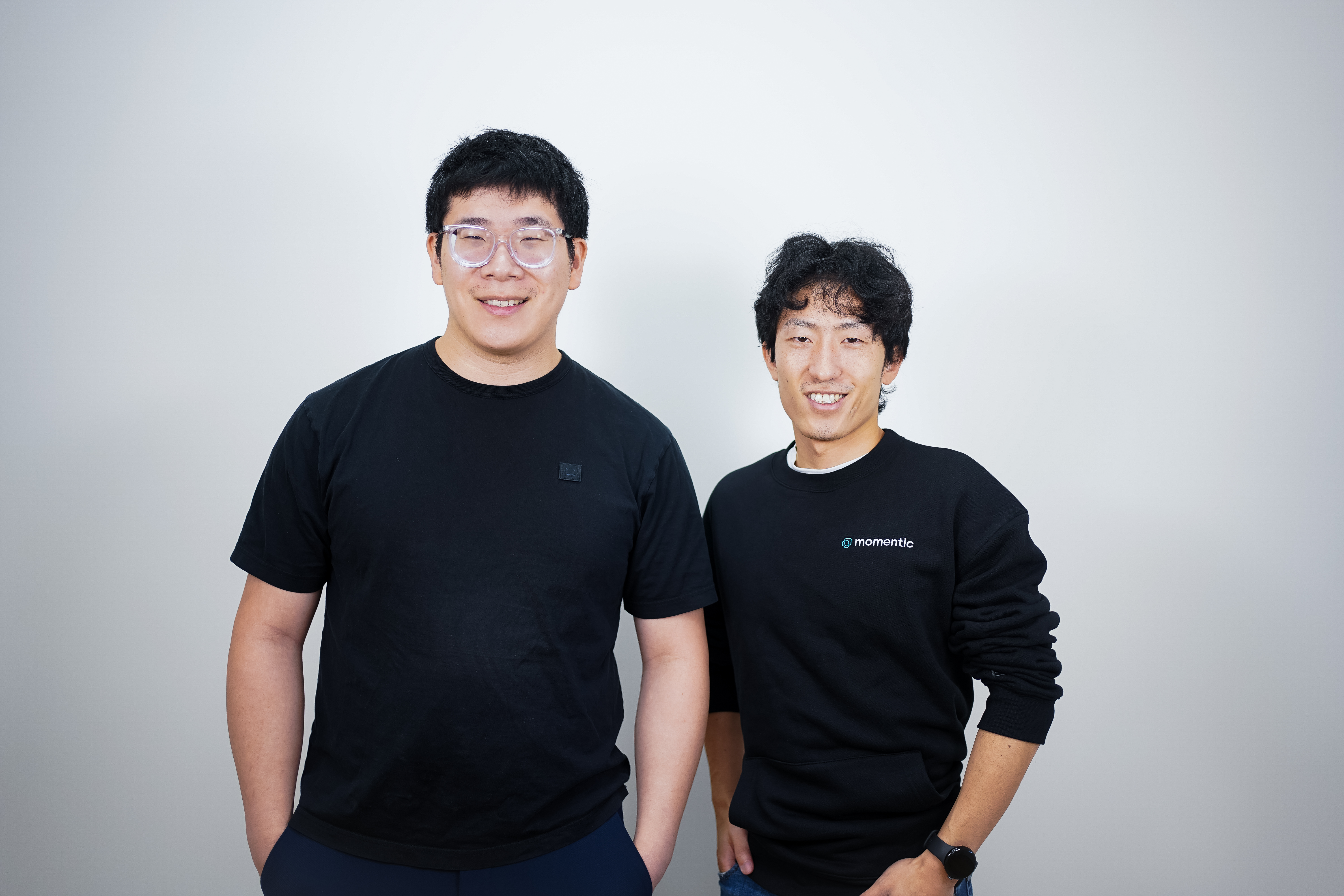 Two men in black t-shirts stand against a gray background.