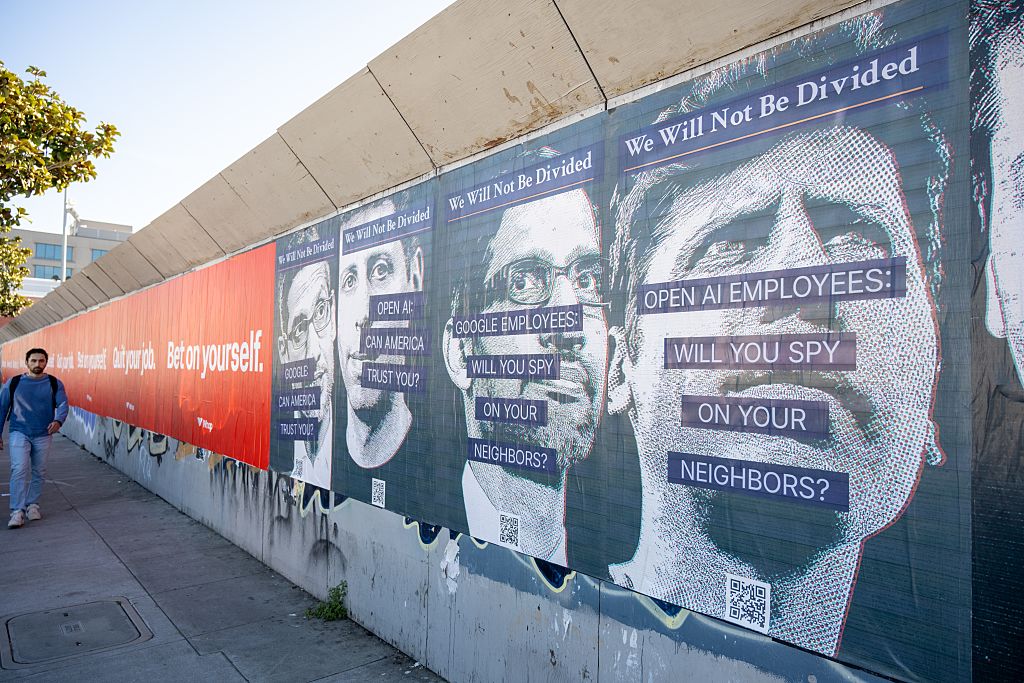 A man walks past a street protest mural referencing OpenAI and Google's artificial intelligence technology and text reading Will You Spy on Your Neighbors, with stylized headshots of the companies' leaders, a reference to controversy regarding artificial intelligence and alleged domestic surveillance, San Francisco, California.