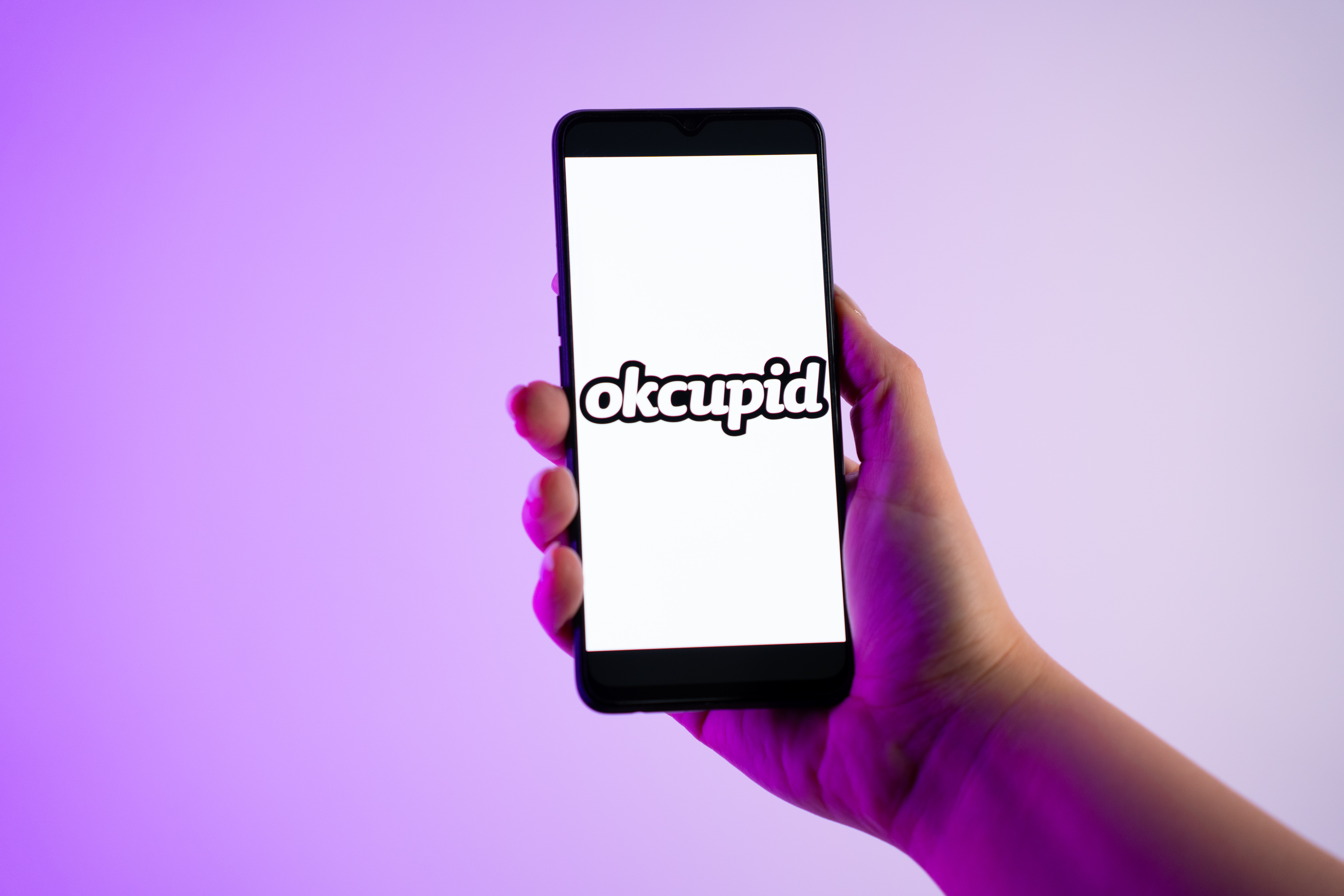 A person is holding a mobile phone with the OkCupid dating app logo on its screen.
