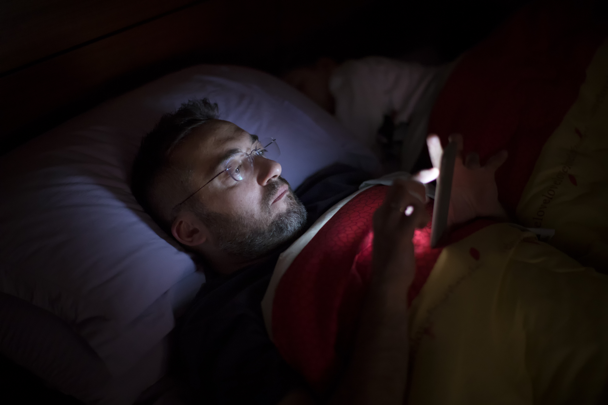 A man in glasses lies in bed, illuminated by the soft glow of his smartphone screen.