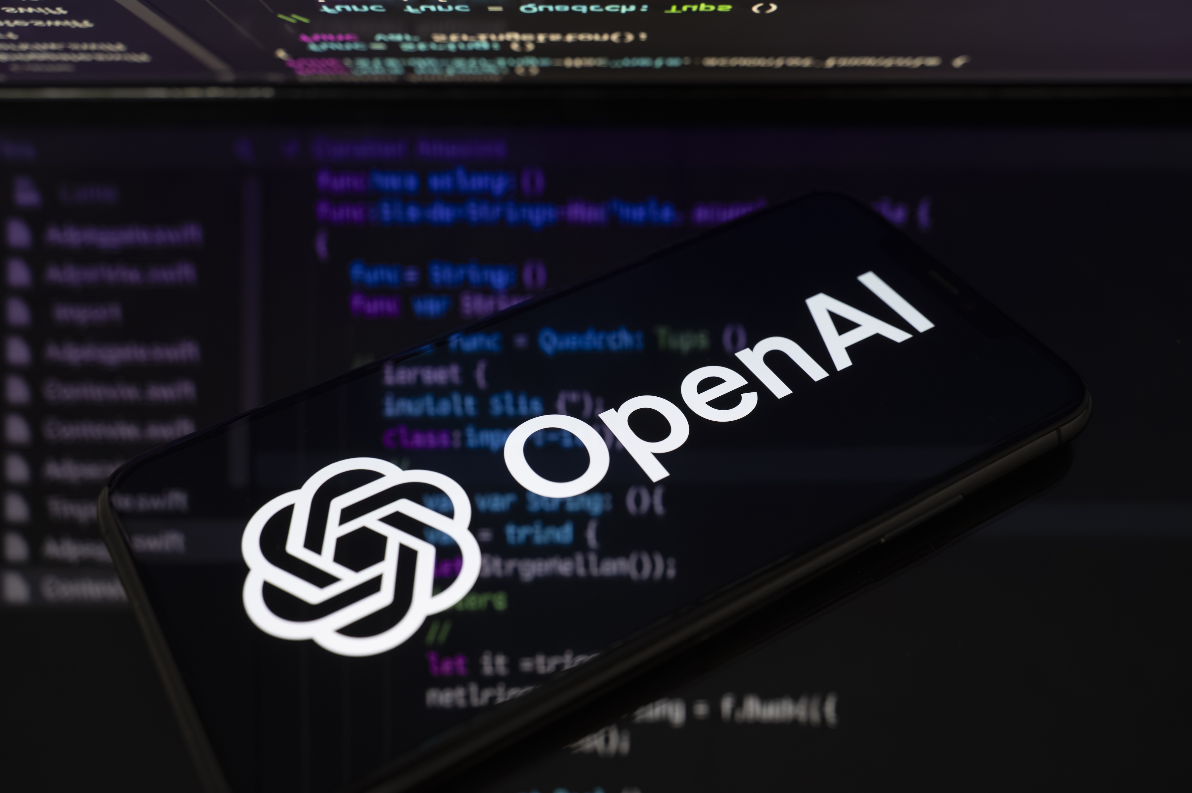 The OpenAI logo is displayed on a smartphone screen placed on a reflective surface onto which lines of computer code.