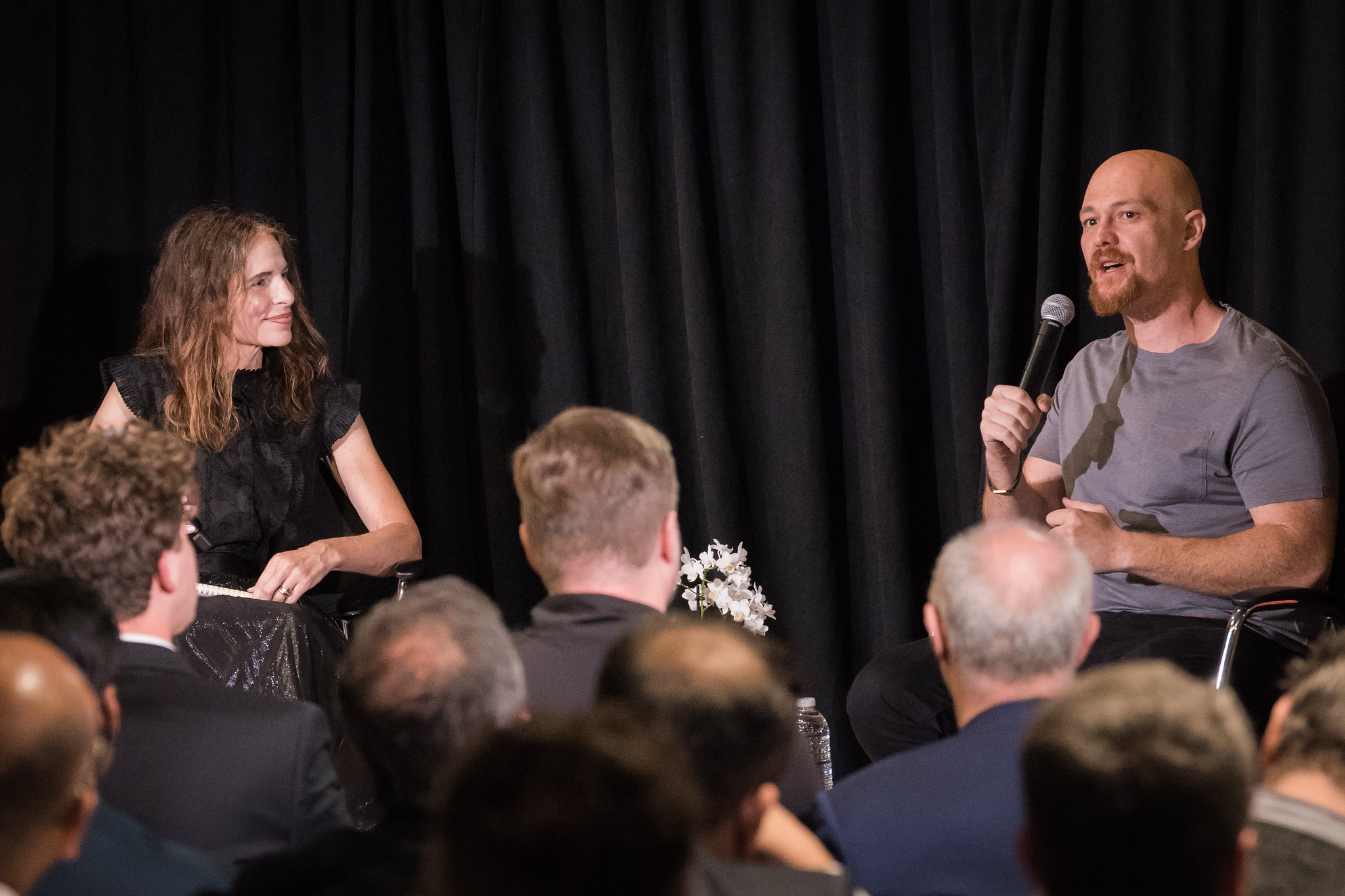 A speaker with a microphone addresses an audience from a stage while seated beside an interviewer. The interviewer, wearing a black outfit, looks toward the speaker as they converse in front of a black curtain backdrop. Audience members fill the foreground, suggesting a live discussion or conference session.