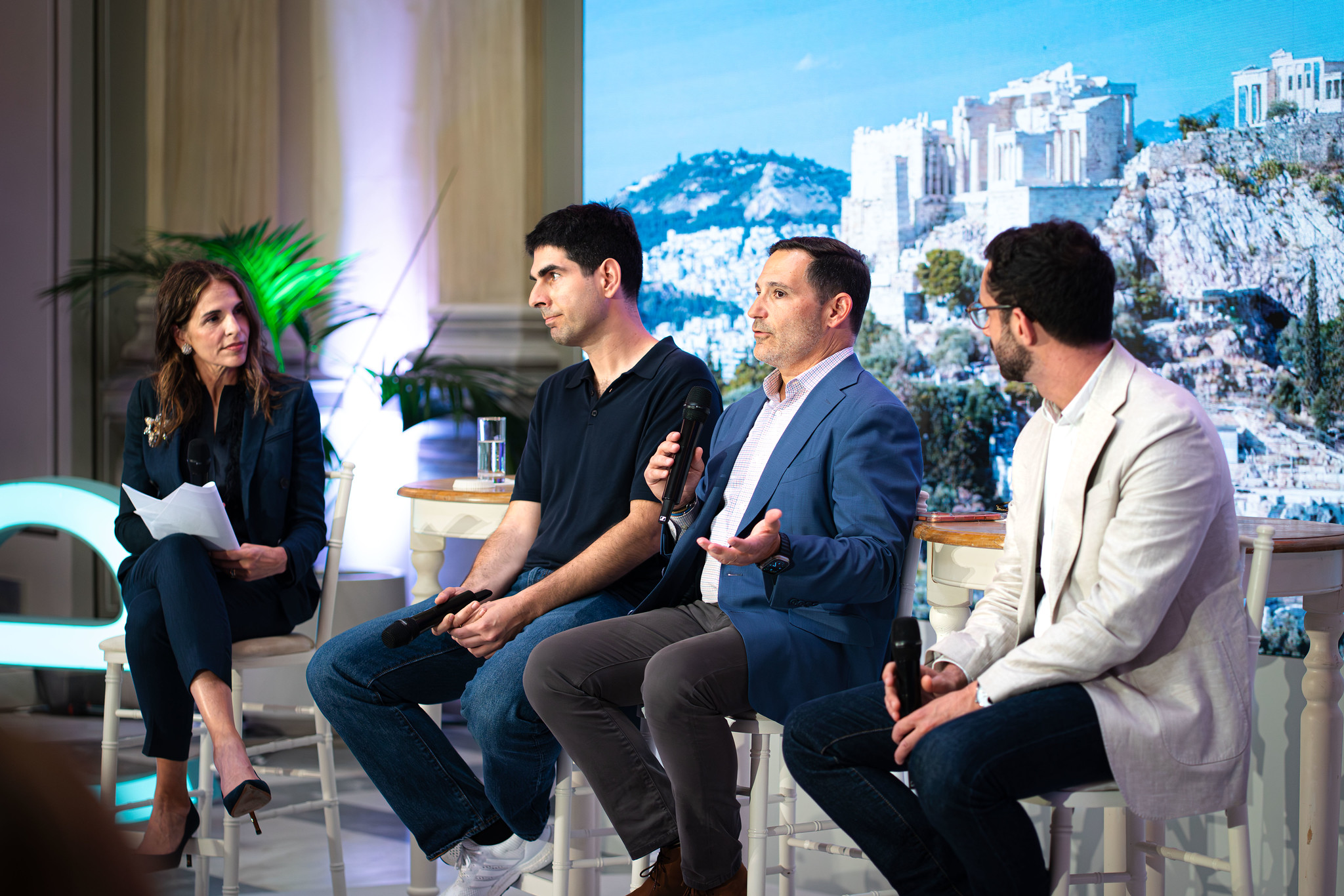 A panel discussion on a stage featuring four seated speakers holding microphones. A woman moderator in a dark suit sits on the left, listening as a man in a blue blazer speaks. Two other men sit beside them, one in a dark polo shirt and one in a light-colored blazer. Behind the panel is a large screen displaying a scenic image of ancient stone ruins on a hillside. Small side tables, glasses of water, and decorative lighting complete the conference setting.