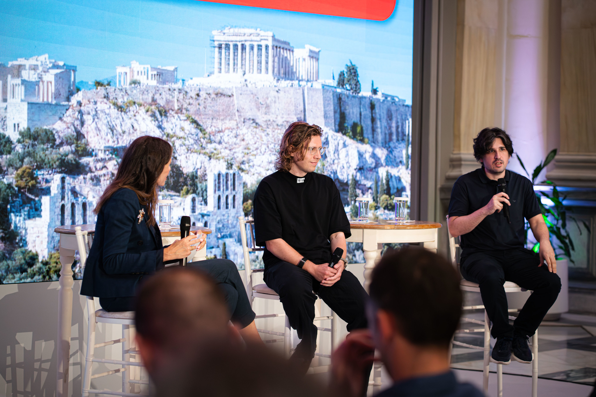 A panel discussion takes place on a stage in front of a large screen displaying the Acropolis in Athens. Three speakers sit on tall white chairs holding microphones, with the person on the right speaking while the others listen. Small tables with glasses of water sit between them, and audience members are visible out of focus in the foreground.