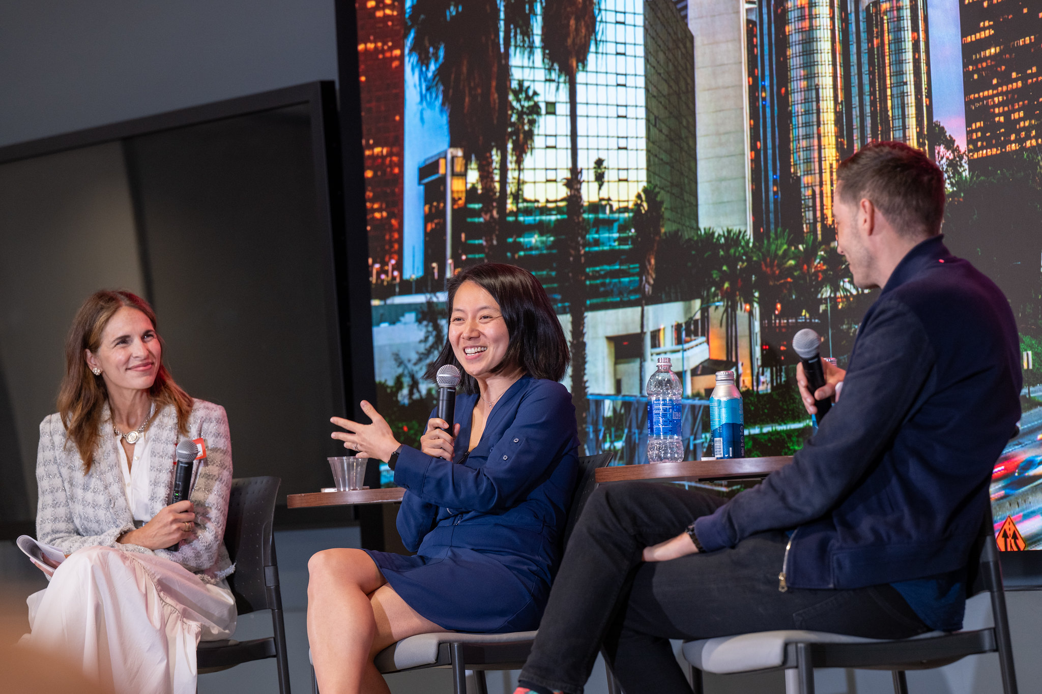 Connie Loizos moderates a panel discussion at StrictlyVC LA, seated on stage with venture capitalists Cheng Xu of Basis Set Ventures and Carter Reum of M13. Xu speaks into a microphone while gesturing, as Loizos looks on and Reum listens. The speakers sit in front of a large backdrop featuring the Los Angeles skyline. Water bottles and microphones rest on small tables between them.
