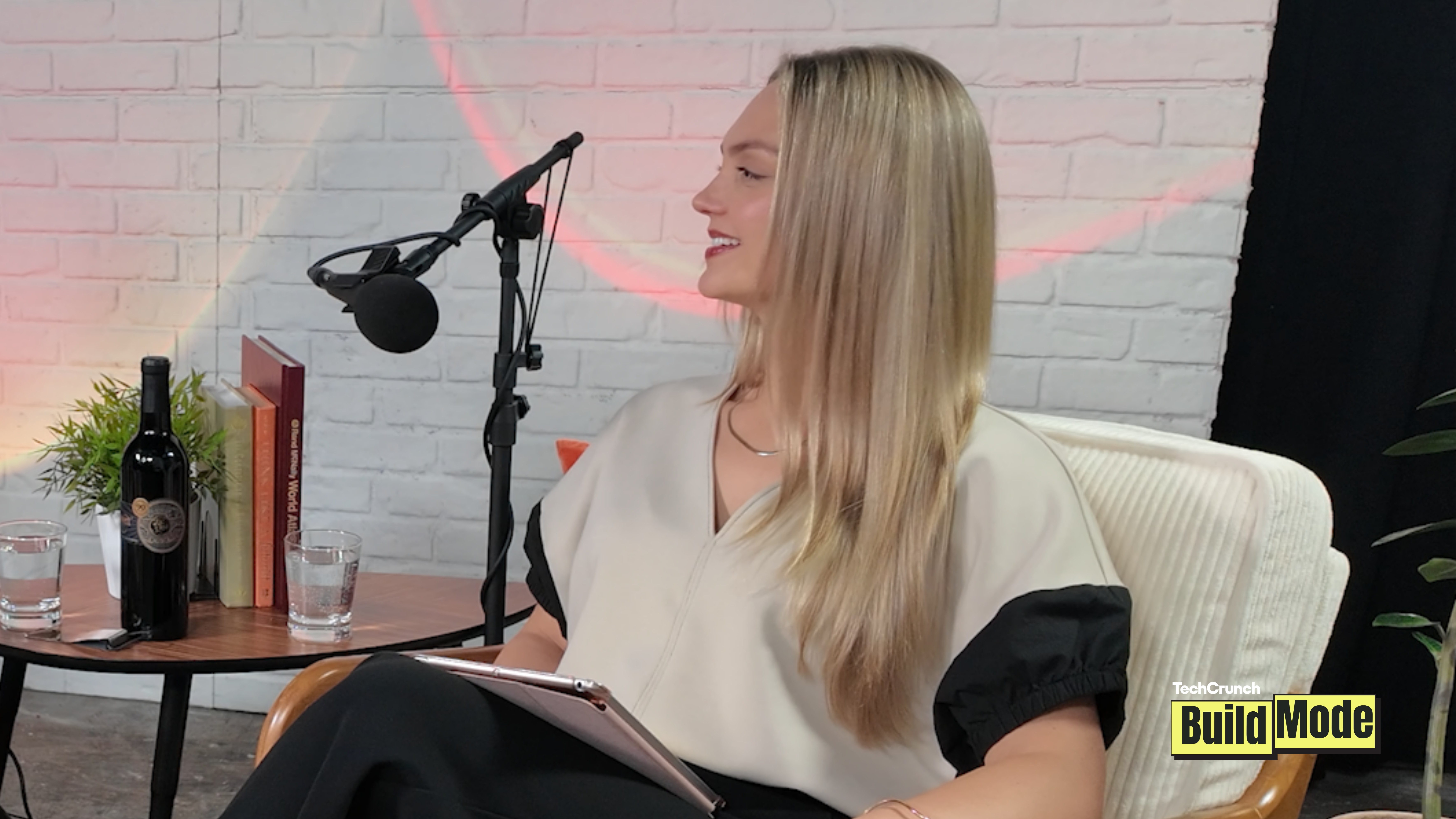 A woman with long blonde hair sits in a white chair during a podcast recording, smiling and looking toward an off-camera guest. A microphone on a boom arm is positioned in front of her, and she holds a tablet on her lap. The studio features a white brick wall backdrop, a small side table with books, glasses of water, a plant, and a decorative bottle. A TechCrunch Build Mode logo appears in the bottom-right corner.